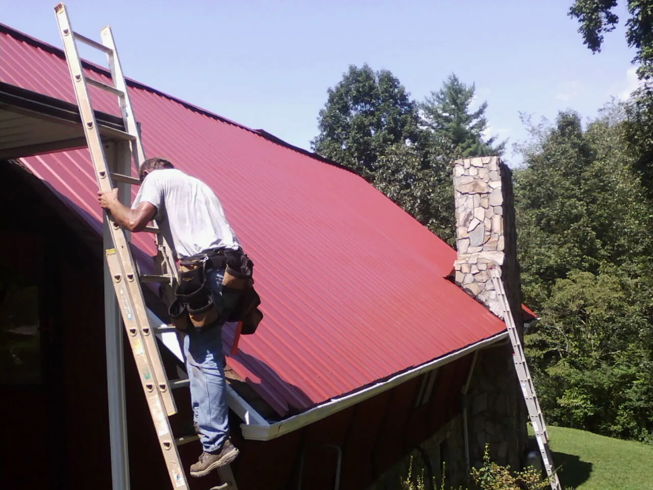 Roofer on a ladder beside a red metal roof and stone chimney — Soffit & Fascia Repair by Roofing Brothers in Glassboro